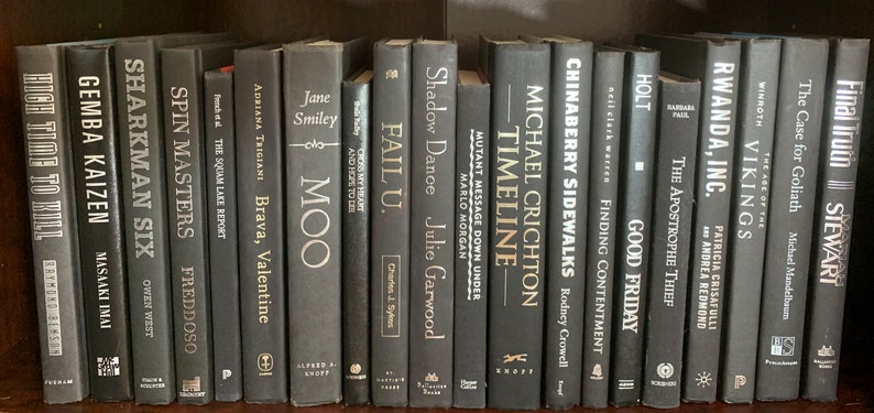 Puede incluir: Una estanter&iacute;a llena de libros negros y blancos con letras doradas. Los t&iacute;tulos de los libros incluyen "Gemba Kaizen", "Spin Masters", "Fail U", "Timeline", "Chinaberry Sidewalks", "Rwanda, Inc.", "The Case for Goliath" y "Final Truth".
