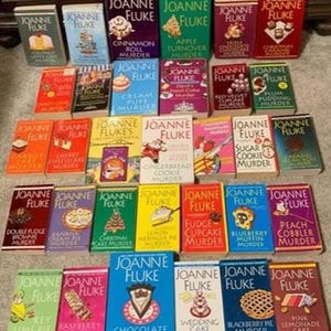 May include: A collection of Joanne Fluke mystery novels, featuring colorful covers with titles like "Cinnamon Roll Murder" and "Chocolate Cream Pie Murder." The books are arranged on a surface, showcasing a variety of dessert-themed mysteries.