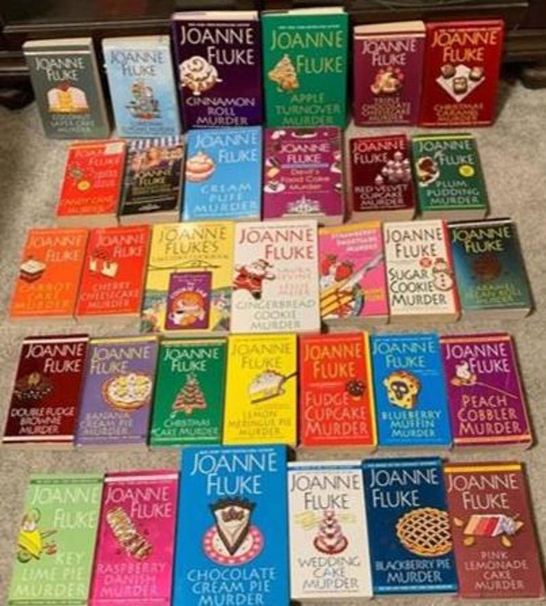 May include: A collection of Joanne Fluke mystery novels, featuring colorful covers with titles like "Cinnamon Roll Murder" and "Chocolate Cream Pie Murder." The books are arranged on a surface, showcasing a variety of dessert-themed mysteries.