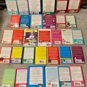May include: A collection of cookbooks with various colored covers, including red, blue, green, and yellow. The books are arranged on a neutral surface, showcasing their diverse titles and designs. Some books feature images of food.