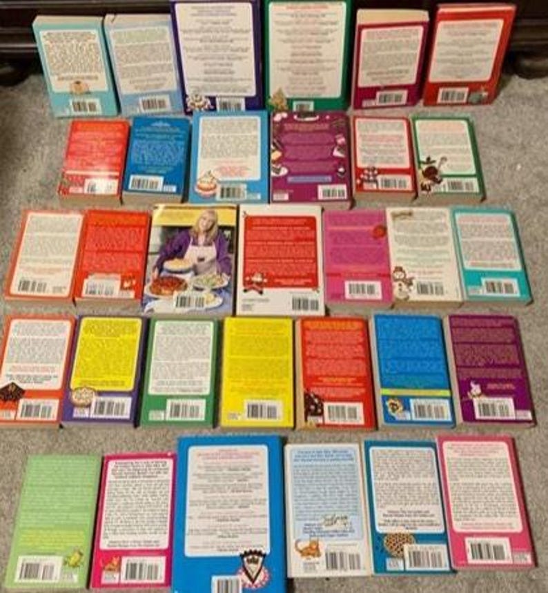 May include: A collection of cookbooks with various colored covers, including red, blue, green, and yellow. The books are arranged on a neutral surface, showcasing their diverse titles and designs. Some books feature images of food.