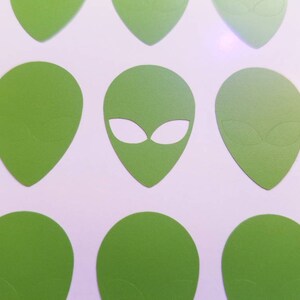 May include: A collection of green alien head cutouts with white eyes. The cutouts are arranged in a pattern on a white background.