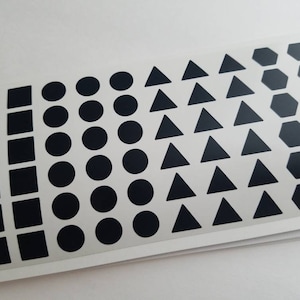 May include: A sheet of black adhesive stickers with various geometric shapes, including squares, circles, triangles, and hexagons.