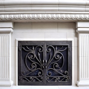 Op de afbeelding: Een witte open haard met een decoratieve lijst boven de mantel en gegroefde kolommen aan beide zijden. Een zwart metalen vuurscherm met een scrollend ontwerp bevindt zich in de opening.