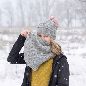 May include: A person wearing a gray knitted hat with a pink pom-pom and a gray knitted scarf with a textured pattern. The person is wearing a black jacket and a yellow sweater. The background is a snowy forest.