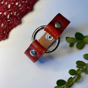 May include: A leather bracelet featuring a stainless steel ring and a snap closure. The leather is a deep reddish-brown. The bracelet is displayed on a white surface, with a red knitted item and green leaves in the background.