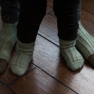 May include: Two pairs of green and brown socks with ribbed knit patterns. The socks are worn on the feet of two people.