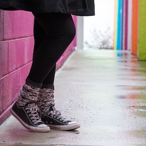 May include: A person wearing black leggings and black and white socks with black Converse sneakers. The person is standing next to a pink wall with a colorful wall in the background.