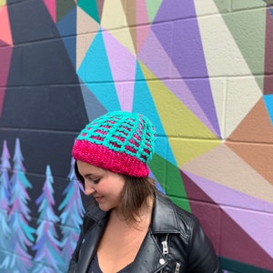 May include: A woman wearing a pink and teal knitted beanie hat with a geometric pattern. She is wearing a black leather jacket and standing in front of a colorful geometric wall.