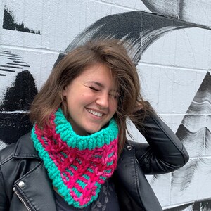 May include: A woman wearing a black leather jacket with a bright pink and teal knitted cowl neck scarf. The scarf is a chunky knit with a geometric pattern.