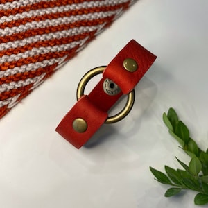 May include: Red leather bracelet with a brass double ring and a small metal tag that says "HANDY".