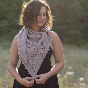 Può includere: Una donna indossa uno scialle di maglia marrone chiaro con un delicato motivo a pizzo. Lo scialle è drappeggiato intorno al collo e alle spalle, e indossa un vestito nero.