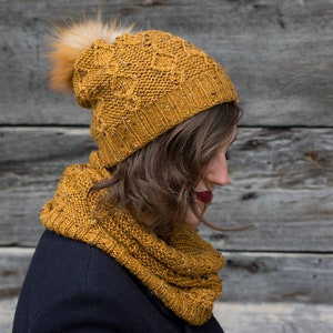 May include: A mustard yellow knitted hat with a fluffy pom pom and matching scarf. The hat and scarf are made with a textured stitch pattern.