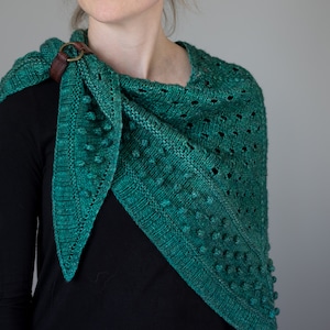 Puede incluir: Un chal de punto verde azulado con un patrón texturizado y una correa de cuero marrón. El chal está drapeado sobre los hombros de una persona.