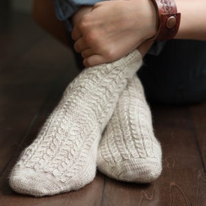 May include: A pair of beige knitted socks with a cable knit pattern. The socks are being worn by a person sitting on a wooden floor.