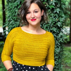 Könnte beinhalten: Eine Frau trägt einen gelben Strickpullover mit einem schwarz-weiß gepunkteten Kleid. Sie steht vor einem grünen, belaubten Hintergrund.