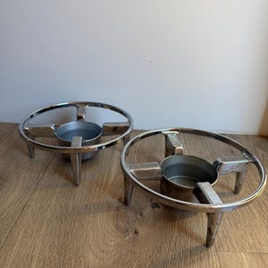 May include: Two circular silver-toned metal stands, each featuring a central cup-like holder supported by three legs. The outer ring is connected to the center by four spokes, with textured rectangular detailing on each spoke.