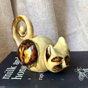 May include: A ceramic cat figurine in cream and brown tones. The cat is curled up with its tail spiraling upwards. The figurine is resting on a book titled "milk and honey".