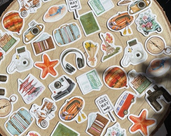 May include: A collection of travel-themed stickers arranged on a wooden surface. The stickers feature illustrations of cameras, luggage, a hot air balloon, a scooter, a passport, a globe, a starfish, and a sign that says "ONE WAY".