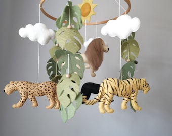 May include: A baby mobile featuring a wooden ring with felt jungle animals, including a leopard, lion, tiger, and black panther. The mobile also includes felt leaves, clouds, and a sun. The mobile is suspended by white strings.