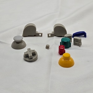 May include: A set of replacement buttons for a gaming console. The set includes a variety of colours, including grey, blue, red, green, and yellow. The buttons are made of plastic and have a variety of shapes, including round, square, and cross-shaped.
