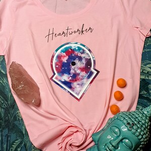 May include: Pink t-shirt featuring a graphic design of a colourful mandala with the text "Heartworker" printed on the front.