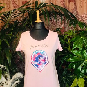 Op de afbeelding: Roze T-shirt met een kleurrijk grafisch ontwerp en de tekst "Heartworker" gedrukt op de voorkant.