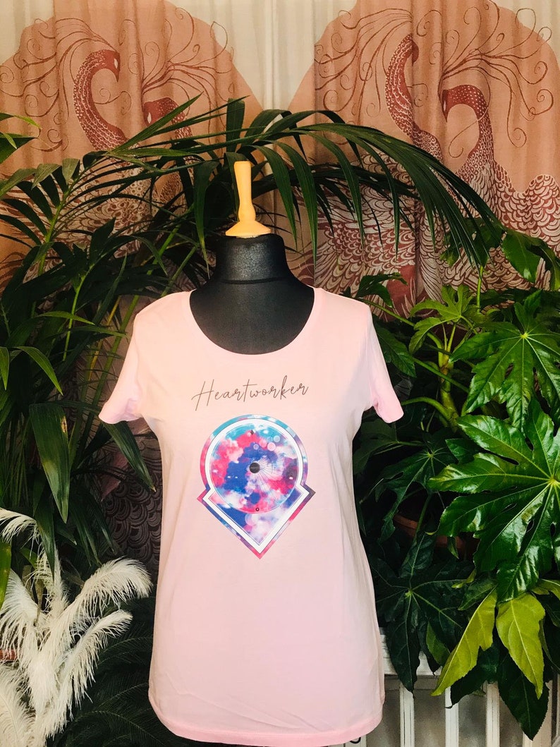 Op de afbeelding: Roze T-shirt met een kleurrijk grafisch ontwerp en de tekst "Heartworker" gedrukt op de voorkant.