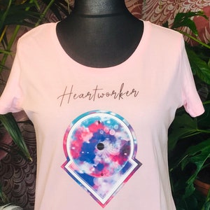Op de afbeelding: Roze T-shirt met een grafisch ontwerp van een cirkel met een blauw, roze en wit tie-dye patroon in een geometrische vorm. De tekst "Heartworker" is in bruin op het shirt gedrukt.