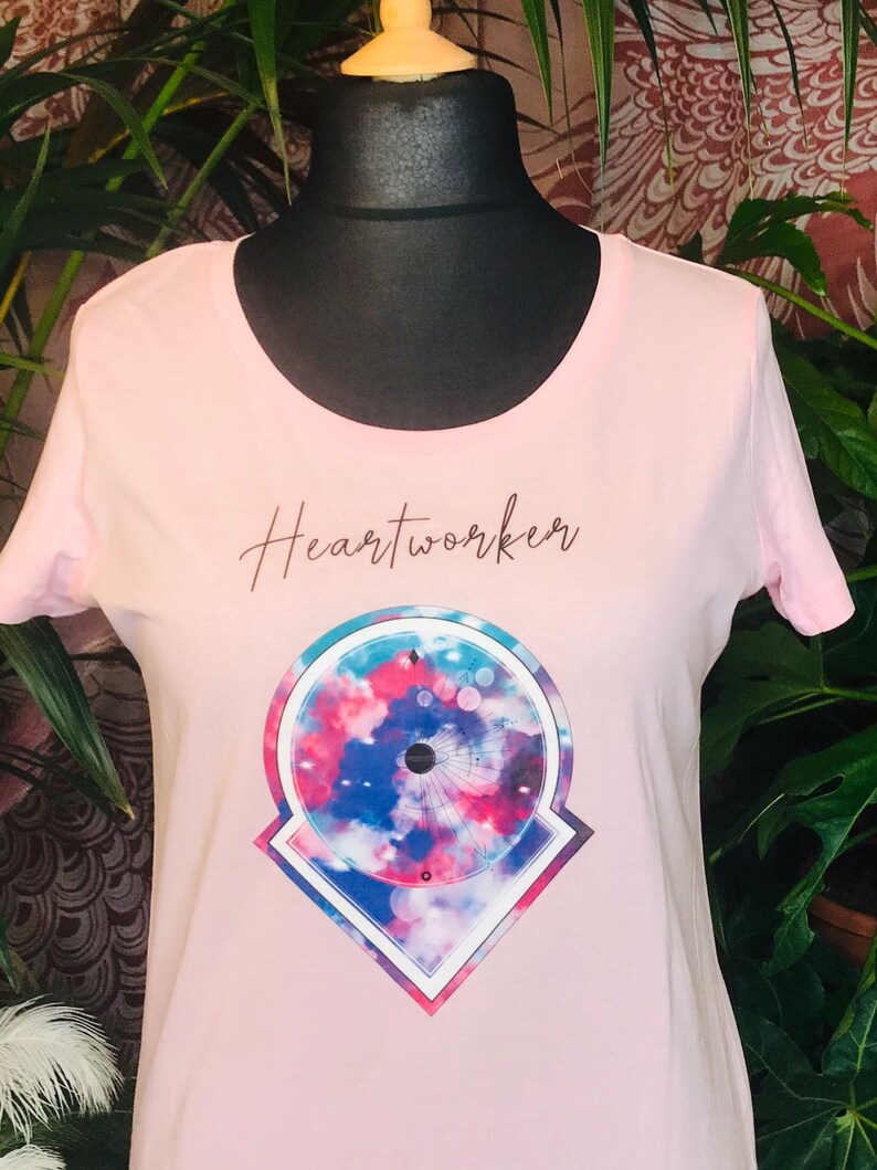 Op de afbeelding: Roze T-shirt met een grafisch ontwerp van een cirkel met een blauw, roze en wit tie-dye patroon in een geometrische vorm. De tekst "Heartworker" is in bruin op het shirt gedrukt.