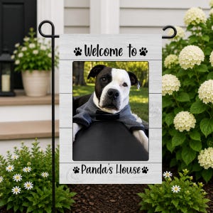 May include: A rectangular garden flag with a white wood-grain pattern featuring a central photograph of a black and white dog wearing a gray shirt. The text "Welcome to" and "Panda's House" is printed in black with paw print accents.