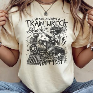 May include: Cream-colored t-shirt featuring a black and white graphic of a skeleton riding a train. The text reads "I'm not always a train wreck, just kidding" and "Toot Toot".