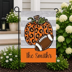 May include: A rectangular garden flag featuring a central orange pumpkin with a black leopard print pattern, a brown football, and the text The Smith's in black script. The background has horizontal gray stripes and a solid orange bottom section.
