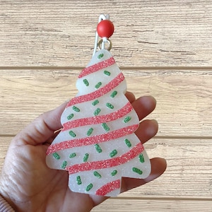 May include: A Christmas tree-shaped ornament with a white base, red glitter stripes, and green sprinkles. The ornament is held by a red bead and a silver-colored metal loop.