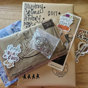 Puede incluir: Un paquete de papel marrón con varias pegatinas y un collar de plata. Las pegatinas incluyen "Travel Time Dude", "You Can Do Hard Things" y "Make Today a Good Day". El paquete está dirigido a "Mystery Crime History Non-Fic 2017*"