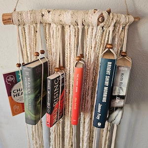 May include: A macrame wall hanging with books hanging from it. The books are titled "Harry Potter and the Half-Blood Prince", "Red Rising", "The Almost Moon", "Bright Shiny Morning", and "Keith Richards Life".