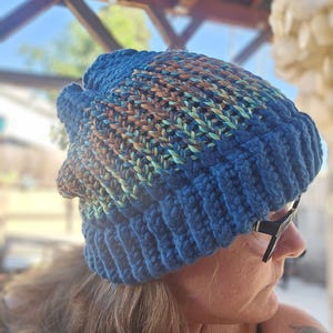 May include: A blue crocheted beanie with a multi-colored stripe pattern. The beanie is made with a chunky yarn and has a slouchy fit.
