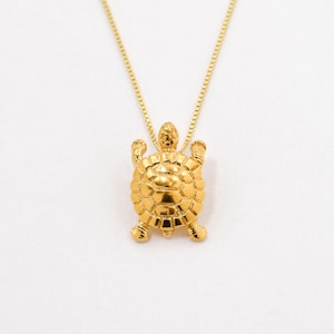 May include: A gold-toned necklace featuring a small, detailed turtle pendant suspended from a delicate box chain. The turtle charm displays a textured shell with defined segments and four visible legs, all rendered in a polished, reflective gold finish.