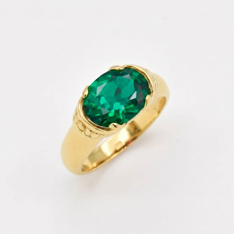 Pu&ograve; includere: Un anello d'oro con una grande gemma verde smeraldo a taglio ovale. La fascia dell'anello &egrave; in oro lucido, con dettagli decorativi vicino all'incastonatura della gemma. La gemma sfaccettata riflette la luce.