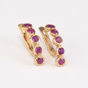 May include: A pair of gold hoop earrings, each featuring four round, deep pinkish-red rubies set in a vertical row along the front curve. The earrings have a hinged closure mechanism and a polished gold finish, showcasing a classic design.