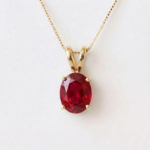 May include: A gold pendant necklace featuring an oval-cut deep red gemstone held by a four-prong setting. The pendant is suspended from a delicate gold chain, showcasing a polished metal finish and a vibrant, saturated crimson stone color.