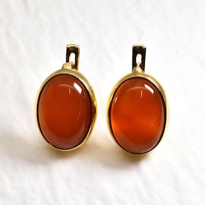 Könnte beinhalten: Ein Paar goldene Ohrringe mit ovalen Karneol-Edelsteinen. Die Ohrringe haben ein schlichtes, klassisches Design.