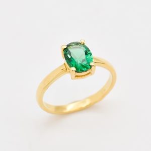 Könnte beinhalten: Ein goldener Ring mit einem leuchtend grünen, oval geschliffenen Edelstein. Der Ring hat ein schlichtes, elegantes Design mit einem schmalen Band und einer Vier-Krappen-Fassung, die den Edelstein sicher hält. Auf schlichtem, weißem Hintergrund.