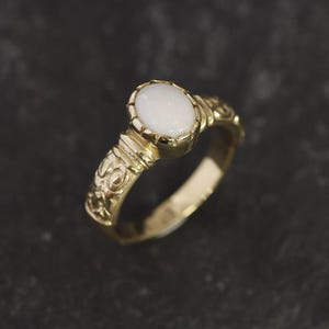 May include: A gold ring with an oval-shaped white opal gemstone. The ring band is decorated with intricate floral designs.