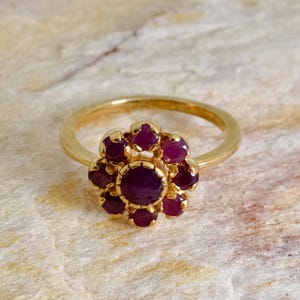 Puede incluir: Anillo de oro con diseño floral. Presenta una piedra central redonda y rojo oscuro, rodeada de pequeñas piedras rojas a juego. La banda es de oro liso. Las piedras tienen un rico color rojo oscuro.