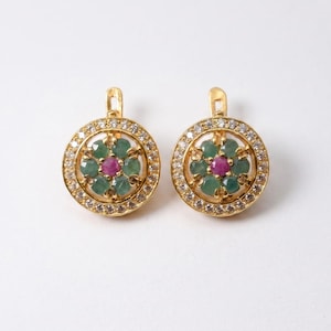 May include: A pair of gold earrings. Each earring has a central ruby surrounded by emeralds, encircled by small clear stones. The earrings have a circular design with a lever back closure. The earrings are approximately 2 cm in diameter.
