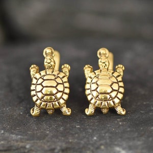 Puede incluir: Un par de pendientes dorados con forma de tortuga. Cada pendiente presenta un diseño detallado de tortuga con caparazón texturizado, cabeza y patas. Los pendientes se presentan sobre una superficie oscura y texturizada.