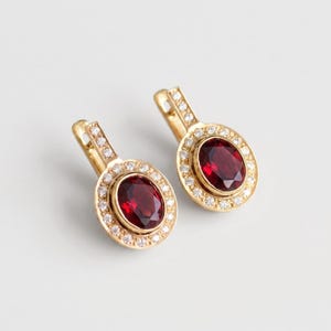 May include: A pair of gold earrings featuring oval-cut deep red garnet stones at the center. Each garnet is surrounded by a halo of small, clear round-cut diamonds. The earrings have a hinged lever-back closure with additional diamonds on the bail.