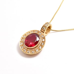 Könnte beinhalten: Eine goldene Halskette mit einem Anhänger, der einen roten ovalen Edelstein mit einem Heiligenschein aus kleineren weißen Edelsteinen zeigt.