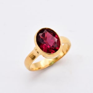 May include: A gold ring featuring a large, oval-cut, deep pinkish-red gemstone set in a smooth, polished gold band. The faceted stone is securely held in a bezel setting, showcasing a vibrant, saturated color and clear, reflective surfaces.